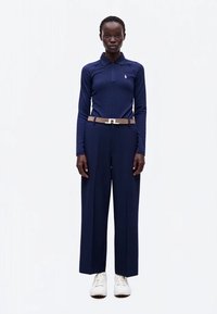 Femme portant un polo bleu marine à manches longues, un pantalon large bleu marine, une ceinture beige et des baskets blanches, debout devant un fond blanc.