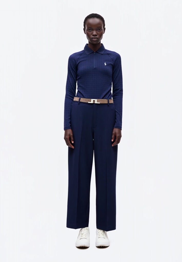 Woman wearing a navy long-sleeve polo shirt, wide-leg navy trousers, beige belt, and white sneakers standing against a white background.