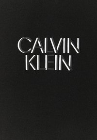 Schwarzer Stoff mit gerippter Textur, der den weißen eingeprägten Text "CALVIN KLEIN" in einer modernen Schriftart prominente zeigt.