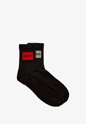 Calcetines negros de algodón con un parche rectangular rojo que lleva el texto "HUGO" en uno y un parche gris con el texto "GO" en el otro. Textura acanalada.