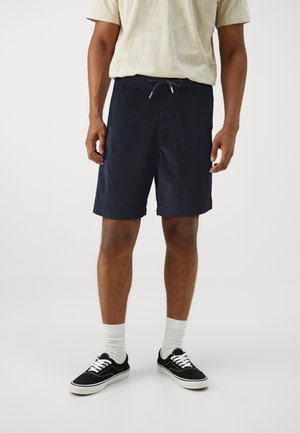 Shorts navy en tissu côte à côte, dotés d'une taille élastique avec un cordon de serrage. Associés à des baskets noires et des chaussettes blanches.