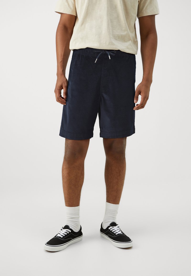 Marineblauwe shorts van corduroy stof, voorzien van een elastische tailleband met een trekkoord. Gecombineerd met zwarte sneakers en witte sokken.