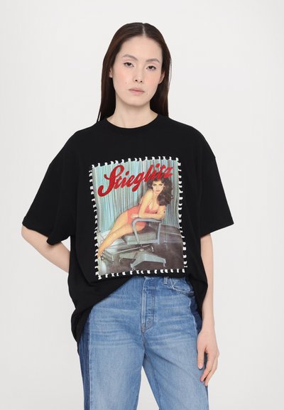 T-shirt oversized negra con un colorido estampado gráfico que muestra a una mujer sentada en una silla, enmarcada por costuras blancas contrastantes.