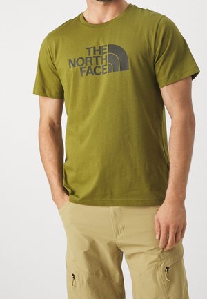 Mężczyzna w oliwkowej koszulce The North Face i beżowych spodniach cargo, z rękami częściowo w kieszeniach na jednolitym tle.