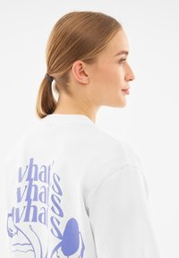 Biała bluza z fioletowym wzorem graficznym na plecach. Tekst i ilustracje zawierają mieszankę organicznych kształtów i wyraźnych liter.