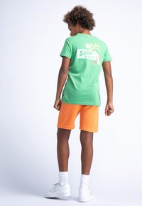 Groene katoenen t-shirt met grafische afbeelding "SURF RIDERS" met palm bomen, gecombineerd met feloranje shorts en witte sneakers. Achteraanzicht.