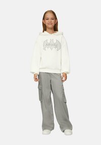 Biała bluza z kapturem z czarnym logo Batmana w kratkę, w połączeniu z szarymi spodniami cargo z bocznymi kieszeniami. Model w białych sneakerach.