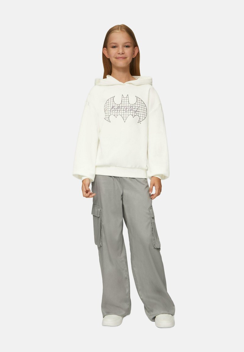 Biała bluza z kapturem z czarnym logo Batmana w kratkę, w połączeniu z szarymi spodniami cargo z bocznymi kieszeniami. Model w białych sneakerach.