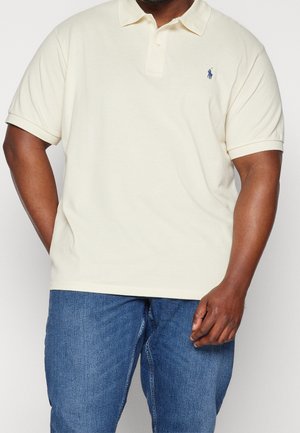 Homme portant un polo à manches courtes crème avec un petit logo bleu sur la poitrine et un jean en denim bleu, debout avec une main dans la poche.