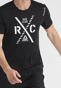 Svart idrottströja med vit grafisk tryck med "RUN CLUB" och "RC". Tillverkad av ett mjukt, andningsbart material med rund hals. Reebok-logga på ärmen.