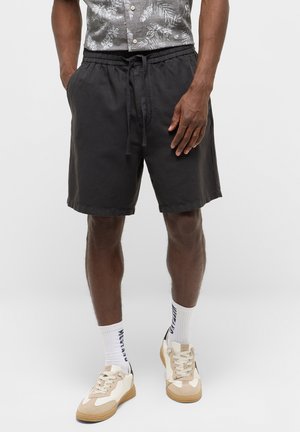 Hombre con pantalones cortos negros con cordón, calcetines blancos altos con texto azul y zapatillas beige y blancas, de pie frente a un fondo liso.