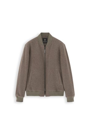 Homme portant une veste bomber grise, une chemise à carreaux, un pantalon noir, des chaussures marron et un bonnet en tricot noir, debout avec une main dans la poche.