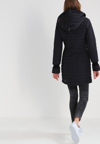Abrigo largo acolchado negro con capucha, que presenta puños acanalados y una cintura ceñida. Tiene una superficie texturizada y un diseño ajustado.