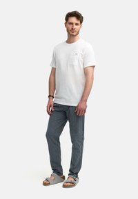 T-shirt blanche à manches courtes avec une poche poitrine, associée à un pantalon gris rayé et des sandales grises avec une sangle et une boucle.