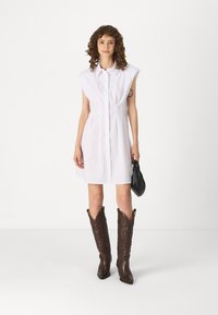 Λευκό, αμάνικο φόρεμα με κουμπί και πτυχώσεις, συνδυασμένο με σκούρα καφέ καουμπόικες μπότες με περίπλοκο ράψιμο. Μαύρη τσάντα χειρός.