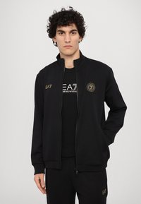 Svart sportjacka med hög krage, dragkedja fram och guld EA7-logga som detaljer. Kombinerad med en svart grafisk t-shirt och matchande byxor.