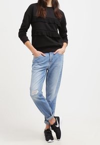 Felpa nera con dettagli a strati in ruffles, abbinata a jeans chiari strappati e sneaker nere. Outfit casual e dal fit rilassato.