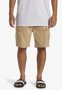 Shorts cargo beiges avec poches latérales, fabriqués en tissu texturé, présentant une coupe décontractée et une longueur au-dessus du genou. Assortis avec des sandales noires.