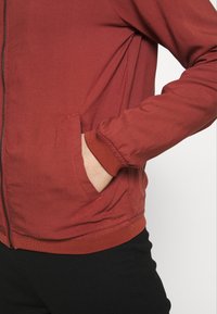 Veste zippée rouge avec poignets et ceinture côtelés, équipée de poches latérales et confectionnée dans un tissu lisse légèrement texturé.