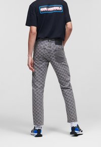 Persona che indossa jeans grigi a fantasia, t-shirt nera con grafica, calzini bianchi e sneakers blu-neri, in piedi su uno sfondo neutro.