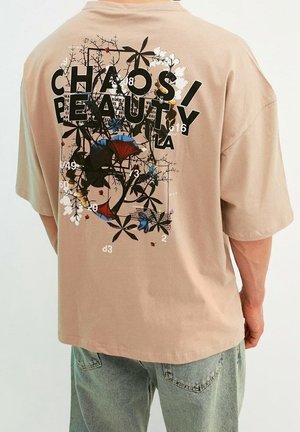 Personne portant un t-shirt oversize beige avec le texte noir « CHAOS BEAUTY » et un graphique floral avec papillon dans le dos, assorti à un jean bleu clair.