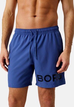 Mannelijk torso met blauwe zwembroek met trekkoord, met "BORG" in het zwart gedrukt op het linkerbeen, staand tegen een effen achtergrond.