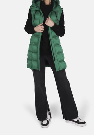 Personne portant une veste sans manches matelassée verte par-dessus un pull noir, un pantalon noir à jambes larges avec des fentes à l'avant, et des baskets noir et blanc.