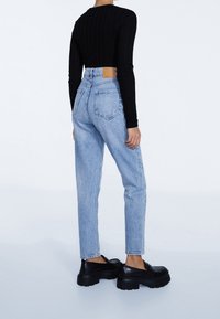Persona vistiendo jeans de tiro alto en azul claro, una blusa negra de manga larga con canalé y unos zapatos de plataforma negros gruesos, de pie contra un fondo blanco liso.