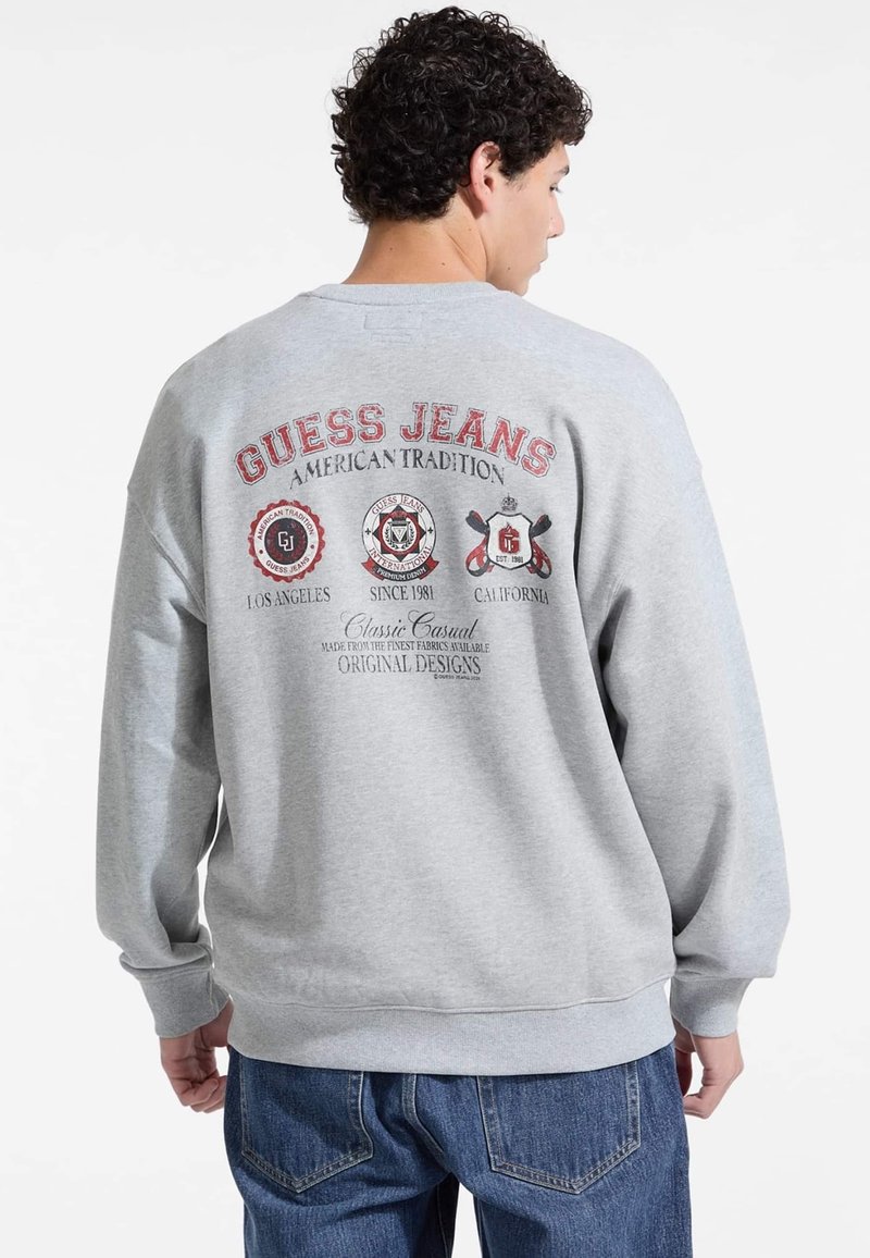 Felpa in cotone grigia con colletto a girocollo, caratterizzata da grafiche stampate sul retro: "GUESS JEANS," loghi e testo in rosso e nero.