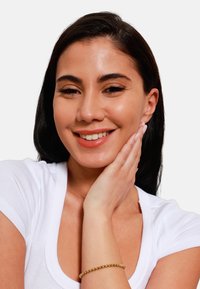 Vrouw met lang donker haar, gekleed in een witte top en een gouden armband. Ze heeft een natuurlijke make-up look en raakt voorzichtig haar gezicht aan.
