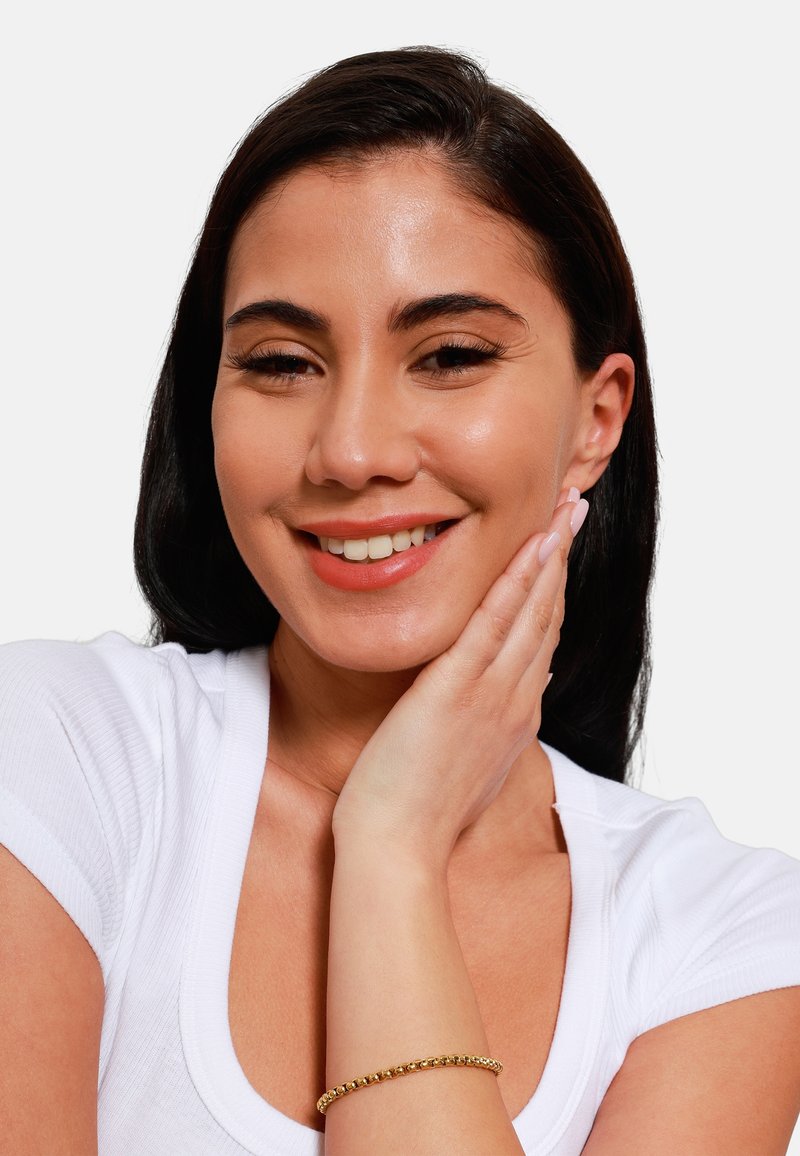 Vrouw met lang donker haar, gekleed in een witte top en een gouden armband. Ze heeft een natuurlijke make-up look en raakt voorzichtig haar gezicht aan.