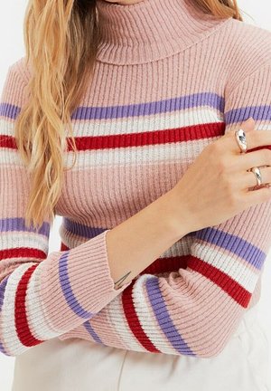 Kobieta w różowym prążkowanym golfie z czerwonymi, białymi i fioletowymi poziomymi paskami, pokazująca srebrne pierścienie i mały tatuaż na przedramieniu.