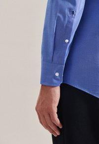 Camicia button-up azzurro chiaro con tessuto testurizzato, dotata di un polsino con un solo bottone e un piccolo dettaglio ricamato su una manica.