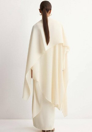 Mujer con coleta larga vestida con un chal estriado en crema de varias capas y falda a juego, de pie contra un fondo claro, de espaldas al espectador.