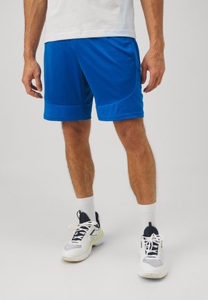 Mężczyzna ubrany w niebieskie krótkie spodenki sportowe, białe skarpetki za kostkę oraz białe i czarne buty sportowe stoi na jednolitym tle.