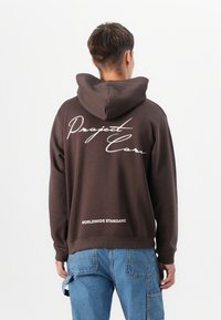 Brown hooded sweatshirt featuring white script text "Project Car" and "WORLDWIDE STANDARD" on the back, with a relaxed fit and textured fabric.