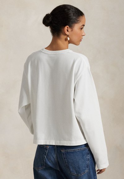 Kvinde iført en løs, hvid langærmet skjorte og blå jeans, håret sat op i en knold, set fra siden mod en neutral baggrund.