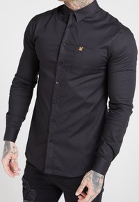 Musta pitkähihainen paita, jossa on napitettava kaulus, rintatasku ja pieni kultainen logo. Sileä kangas ja istuva malli.