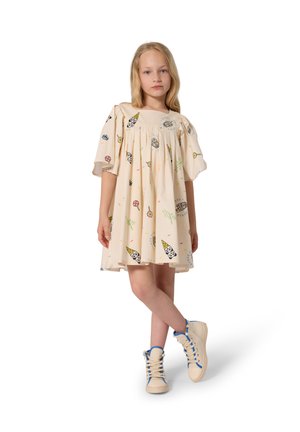 Jeune fille debout, jambes croisées, portant une robe beige clair avec des imprimés de glaces et sucettes en dessin animé, ainsi que des baskets crème avec des accents bleus.