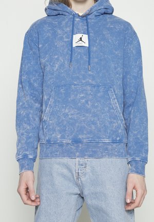 Osoba ubrana w niebieską bluzę z kapturem typu acid-wash z przednią kieszenią kangurka oraz czarnym logo Jumpman na białej naszywce na klatce piersiowej.