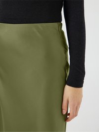 Olijfgroene satijnen rok met een gladde textuur en details in de zijnaad, gedragen met een zwart langemouwtop. Hand ligt lichtjes op de rok.