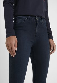 Jeans de mezclilla oscura con una silueta ajustada, con una cintura alta, detalles de costura sutiles y cuatro bolsillos. Combinados con una blusa de manga larga color marina.