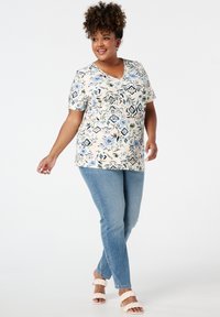 Korte mouwen V-hals top in crèmekleur met een blauw en groen bloemen- en geometrisch patroon; gecombineerd met lichtblauwe slim jeans en gevlochten sandalen.