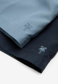 Două perechi de pantaloni scurți: unul albastru deschis, unul bleumarin închis. Ambele au broderie cu palmieri albaștri deschiși aproape de tiv. Textură de țesătură netedă.