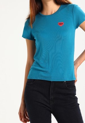 Woman wearing a teal short-sleeve shirt with a small red heart logo reading "OBEY" on the left chest, paired with black pants.