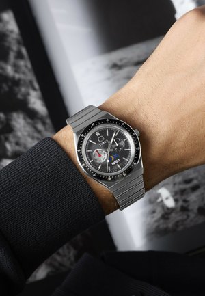 Handgelenk mit silbernem Metallarmbanduhr, die die Zeit 10:10 zeigt, schwarzes Zifferblatt, Unterzifferblätter, NASA-Logo und Mondphasenanzeige an einem dunklen Ärmel.