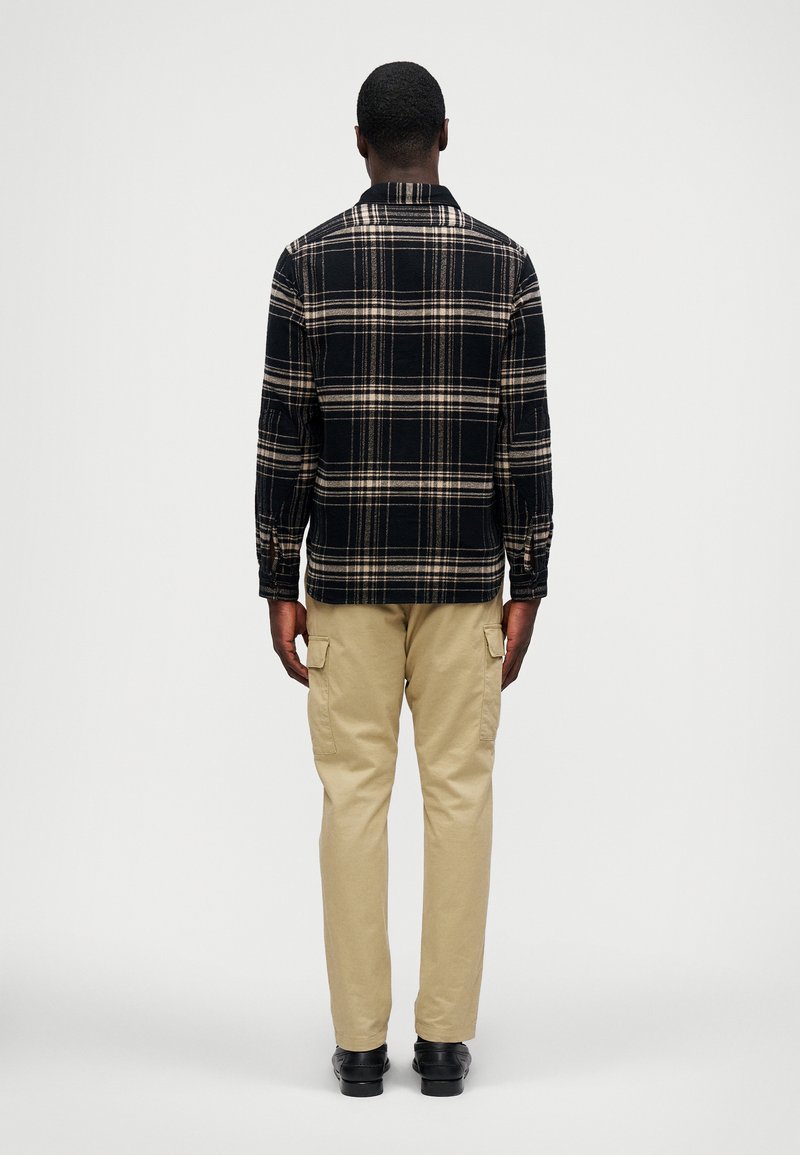 Camisa de cuadros en negro y beige de manga larga con cuello, combinada con pantalones cargo de color beige claro; mostrada desde la parte de atrás con detalles mínimos.