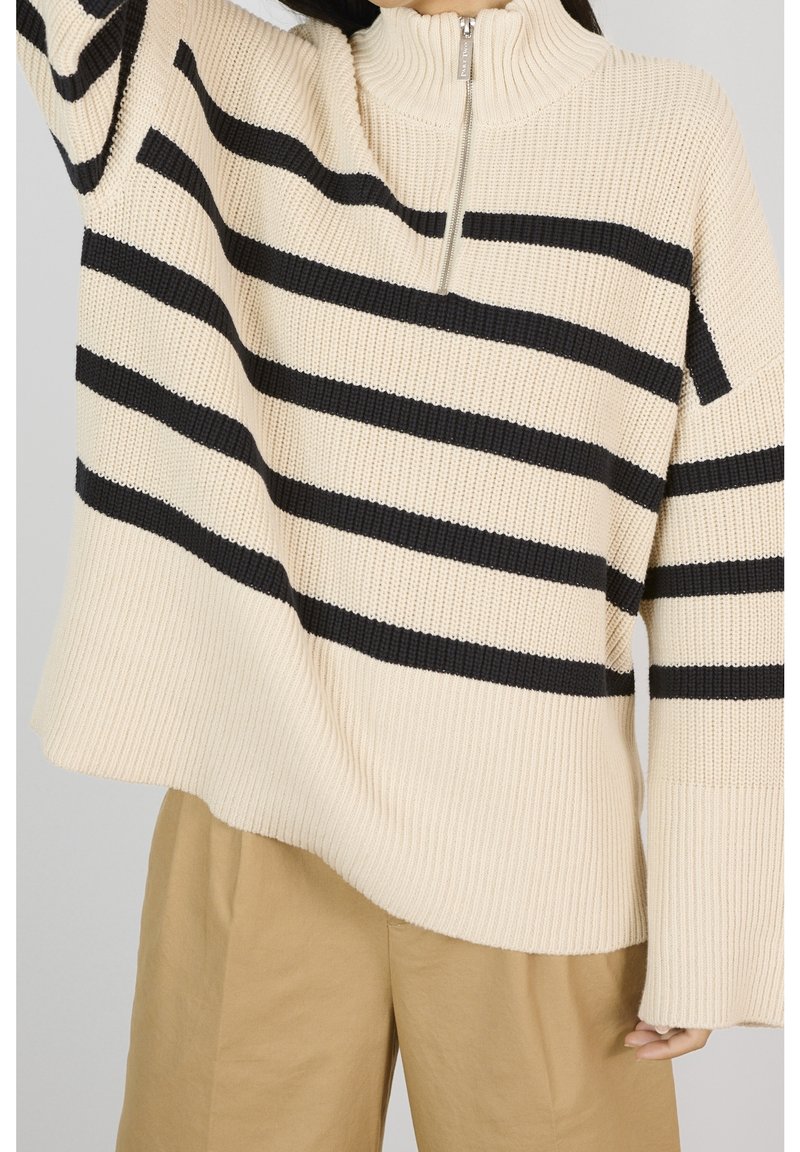 Personne portant un pull côtelé beige à rayures horizontales noires et col zippé à moitié, associée à un pantalon beige ample, devant un fond uni.