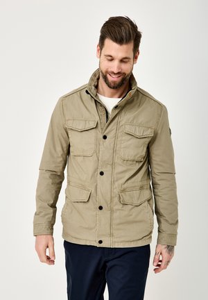 Man with beard smiling, wearing beige jacket with four front pockets and dark pants, standing against a plain light background.