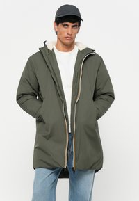 Parka zippé vert olive avec une doublure en polaire blanche texturée. Comprend une capuche, des poches latérales et une fermeture éclair jaune contrastante. Jean en denim porté en dessous.